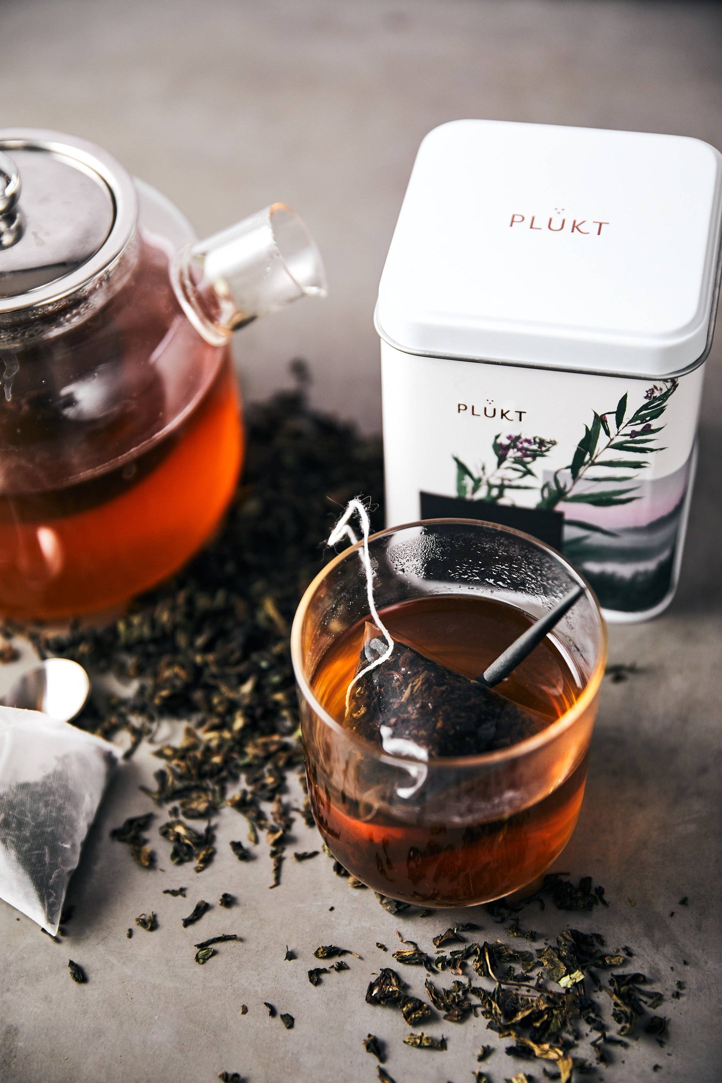 Tea set with glass teapot, cup, and 'PLUKT' tea tin on a wooden surface.
