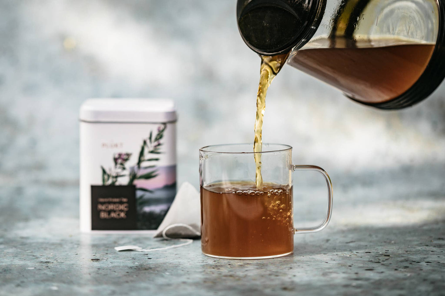 Tea being poured from a pot into a glass mug with a tea canister labeled 'Nordic Black' in the background.