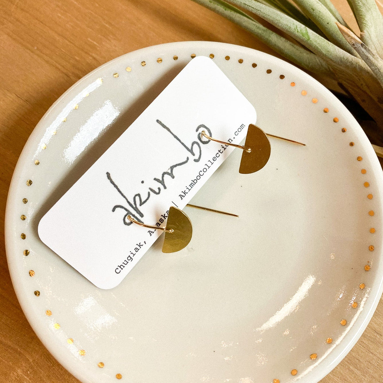 Brass earrings by Akimbo on white ceramic dish with gold polka dots on a wooden surface with a plant.