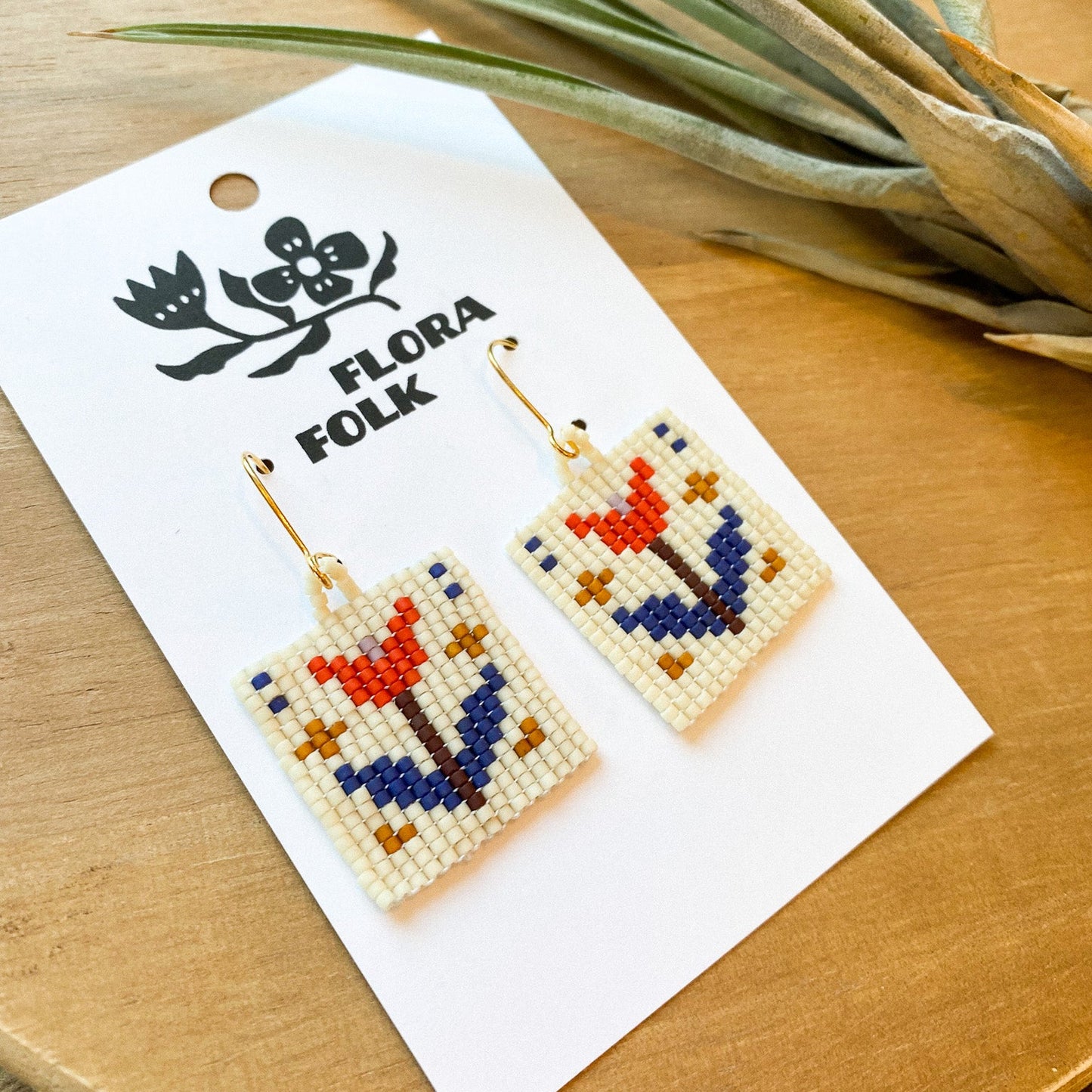 Floral beaded earrings on a 'Flora Folk' branded card with a plant in the background.