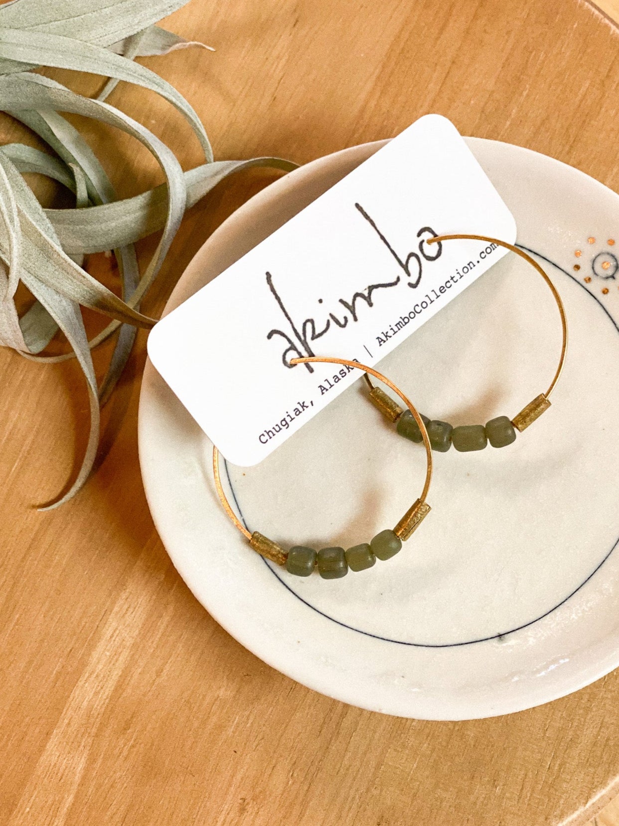 Pair of gold hoop earrings with green beads on a white plate, branded 'Akimbo', on a wooden surface.
