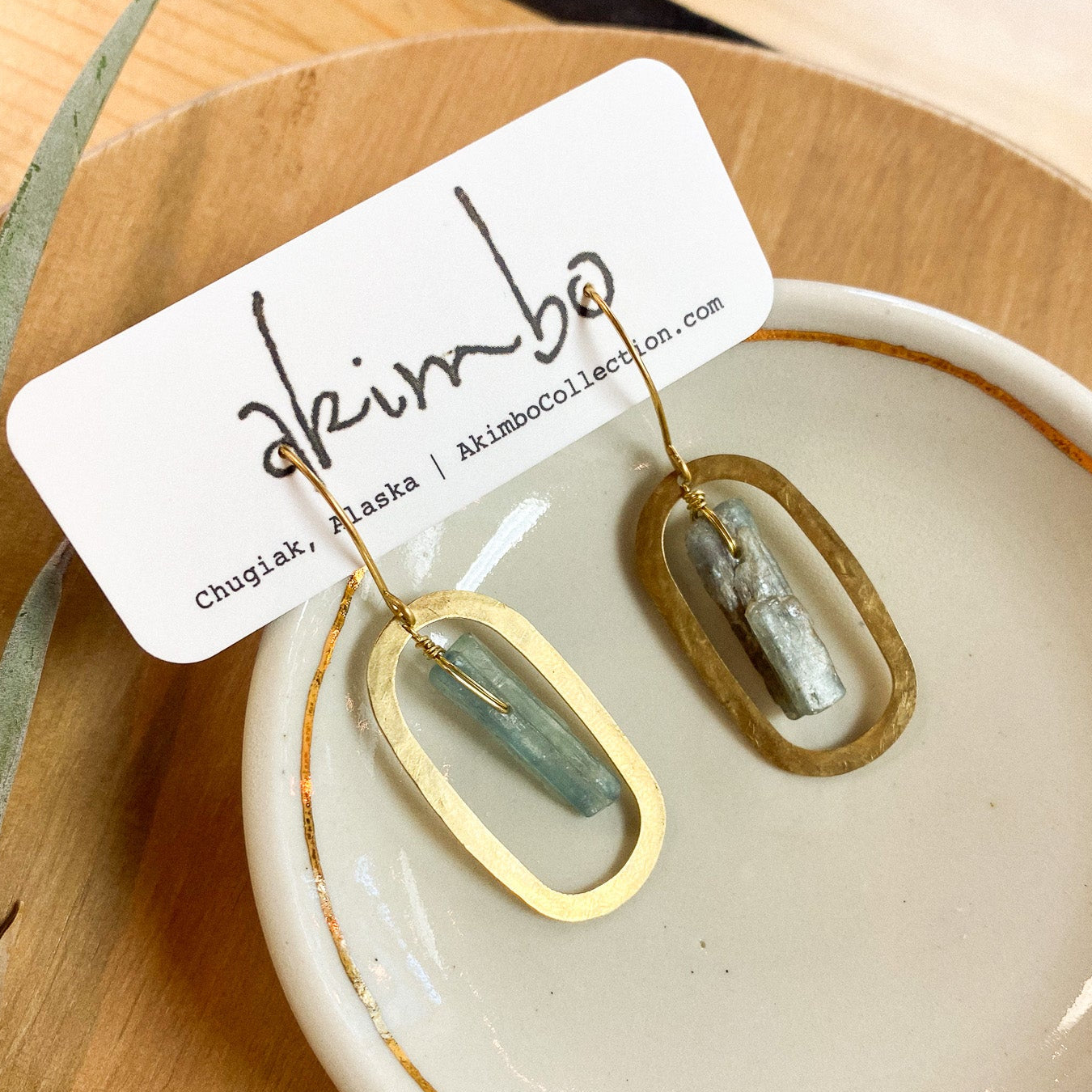 Gold and green earrings on a white dish with a 'akimbo' label on a wooden surface.