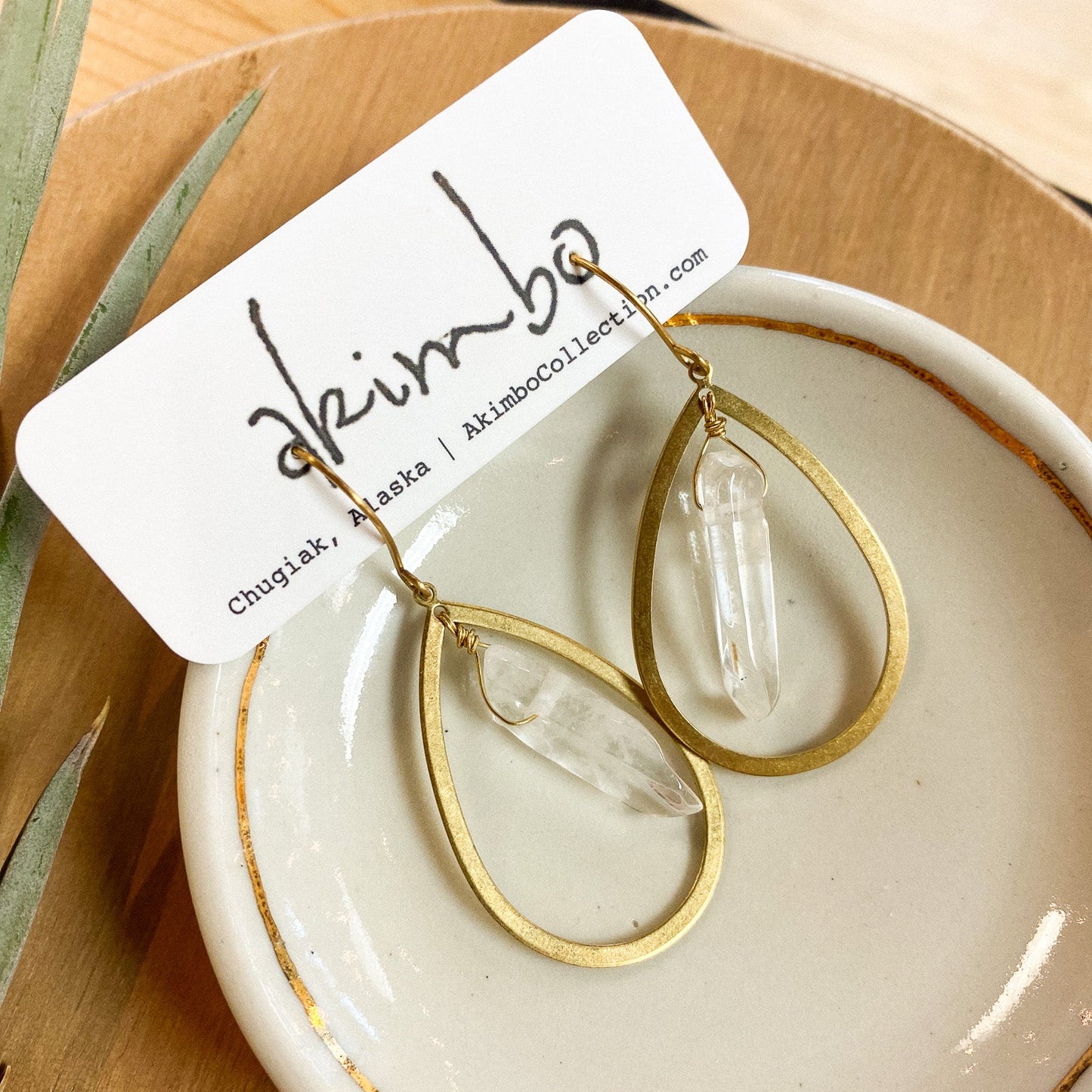 Gold and clear teardrop earrings on a white dish with a branded card, on a wooden surface.