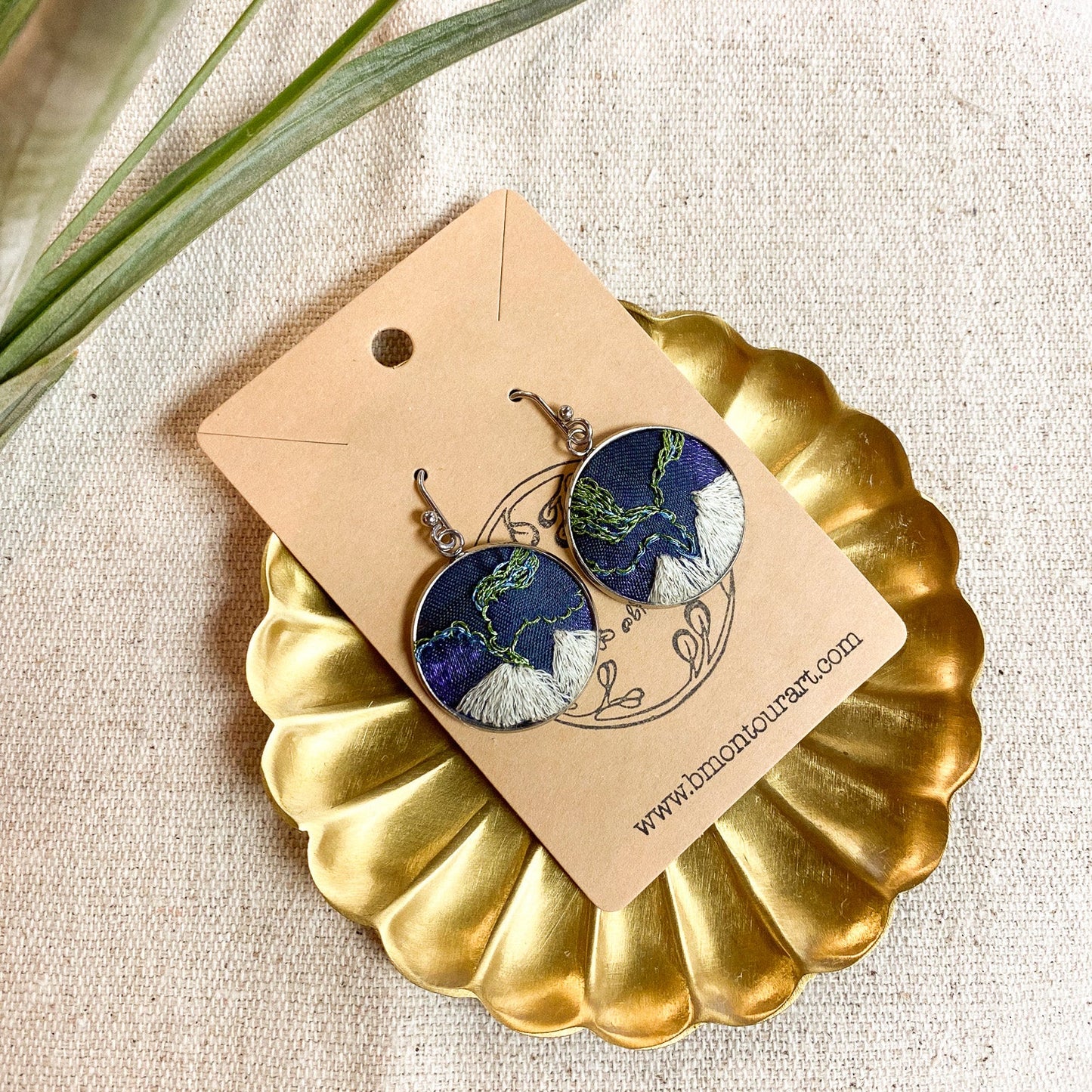 Pair of earrings on a gold display stand with a beige card, on a textured fabric background.