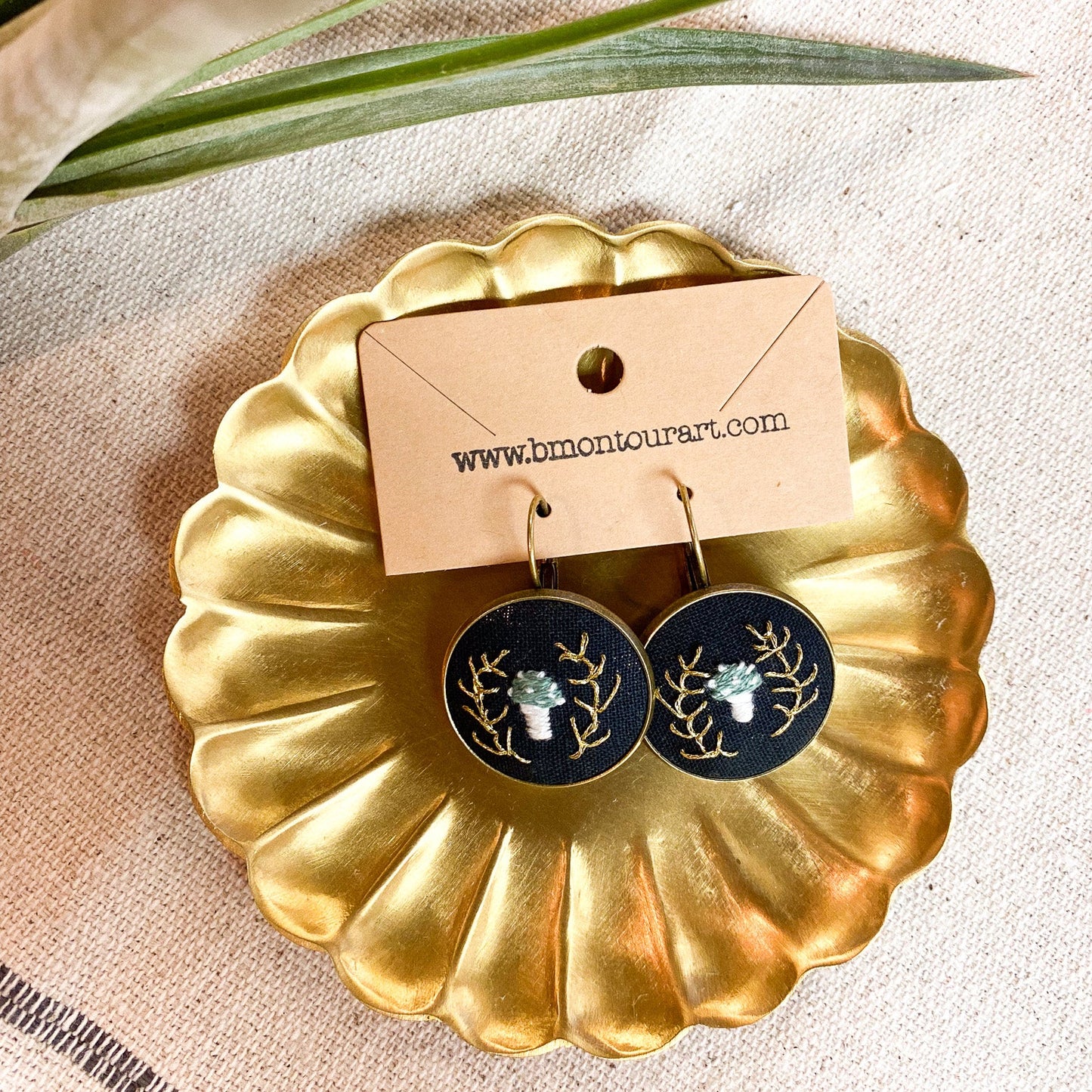 Gold-framed earrings with a nature-themed design on a beige surface with a plant leaf.