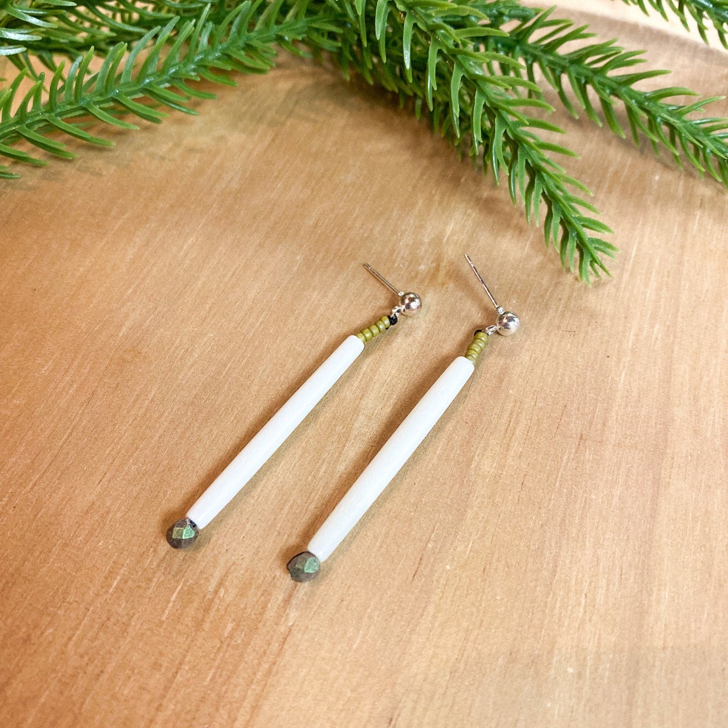 Two white earrings with green accents on a wooden surface, surrounded by green leaves.
