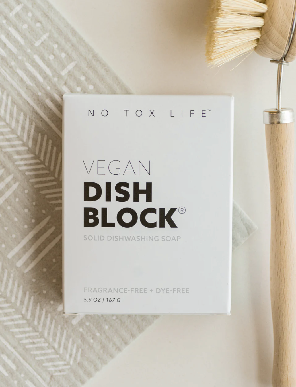 A white dish block soap labeled 'VEGAN DISH BLOCK' by No Tox Life, placed on a patterned surface with a dish brush beside it.