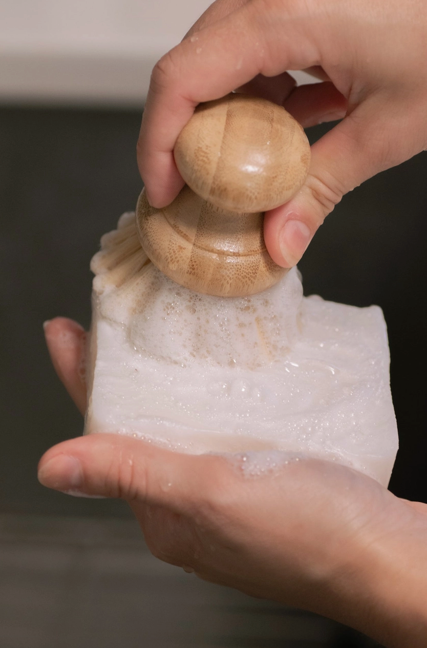 Hand holding bamboo brush and creating suds on a plastic free bar of dish soap which is eco friendly.