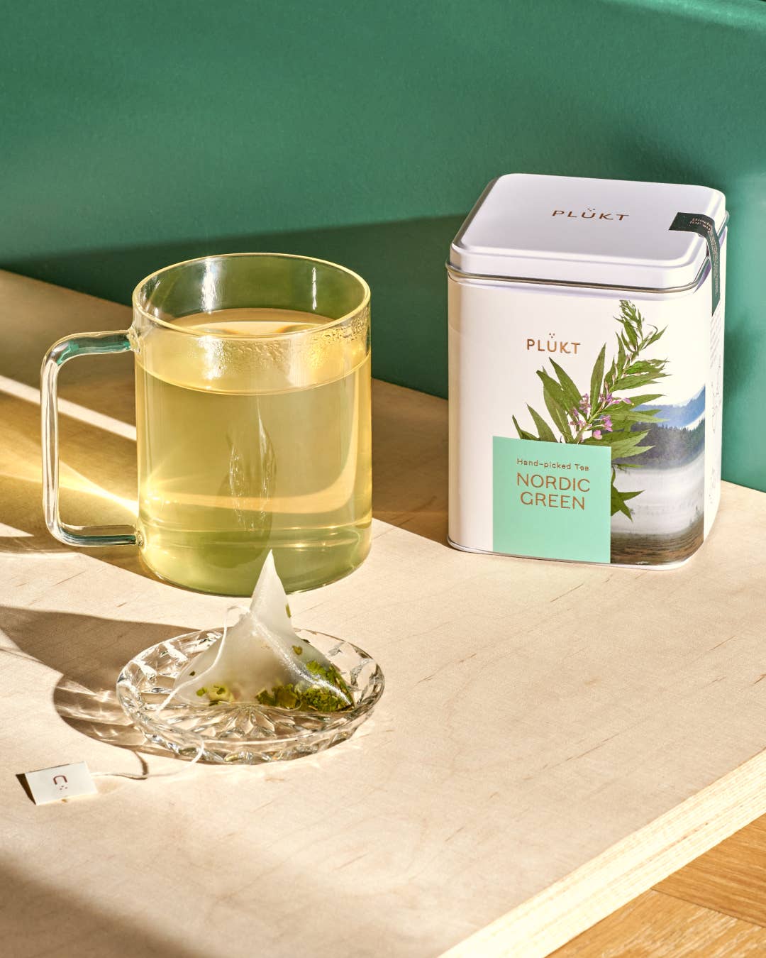 Clear mug with green tea and a teabag, next to a PLUKT Nordic Green tea canister on a wooden surface.
