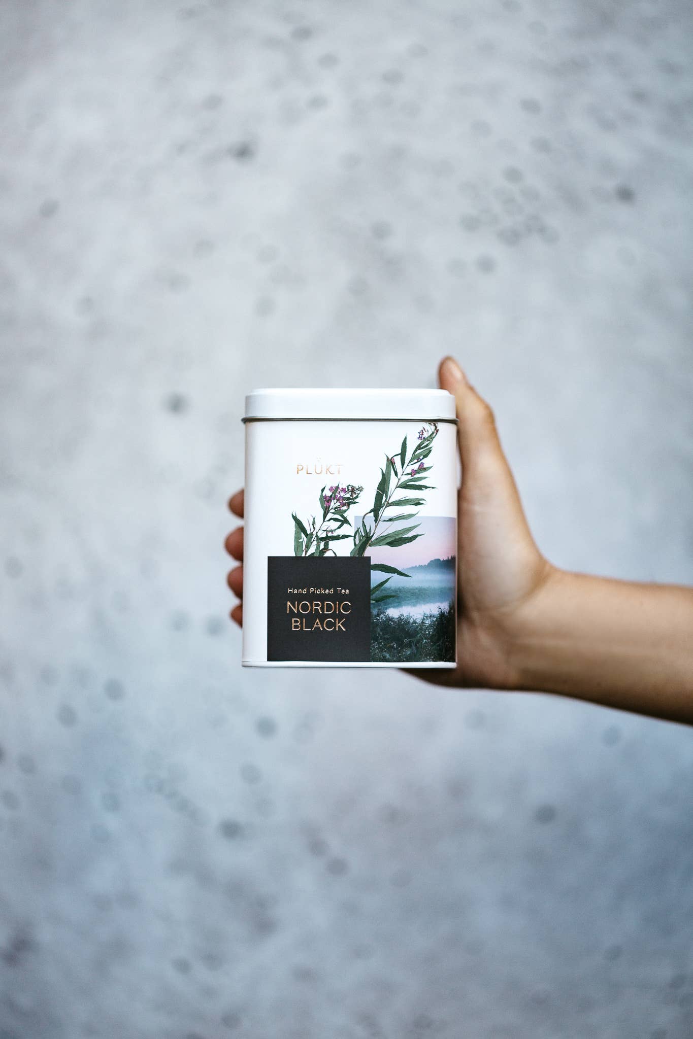 Hand holding a tea canister with a scenic label against a gray background