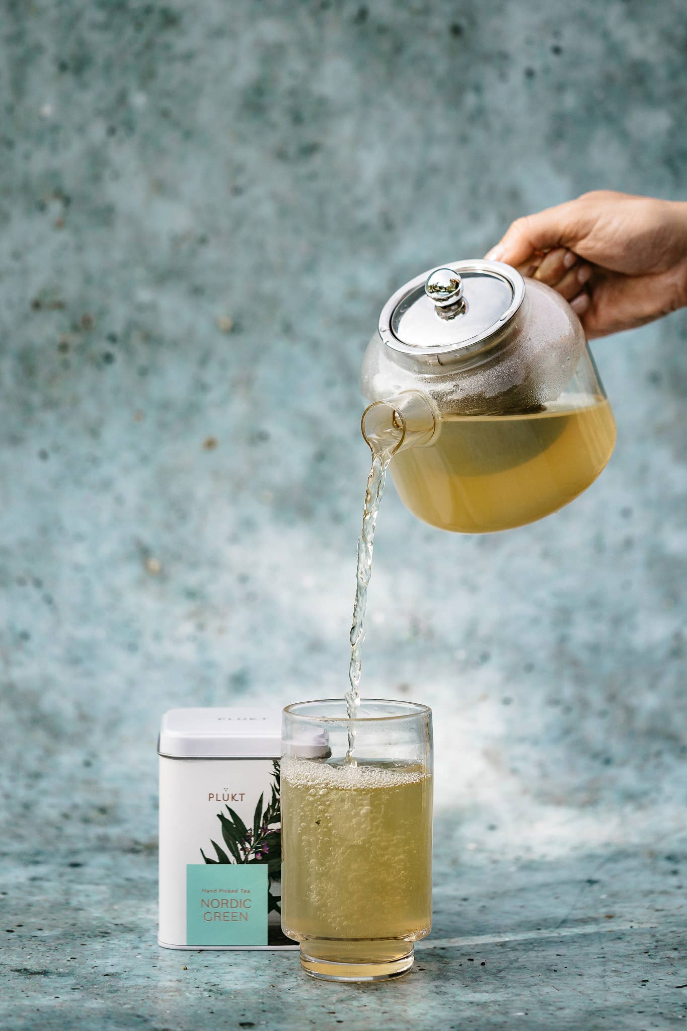 Tea being poured from a glass teapot into a glass on a textured surface with a blurred background