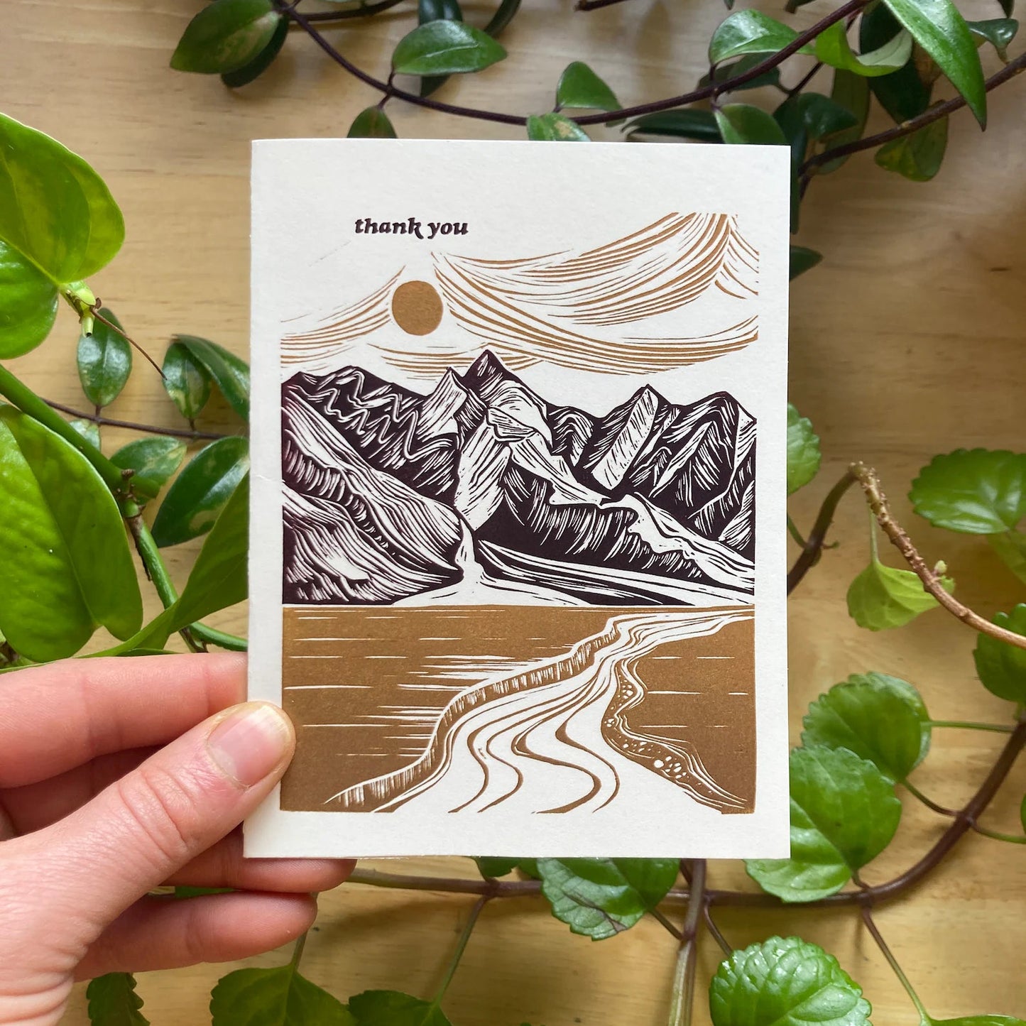 A hand holding a thank you card with a mountain range illustration, accompanied by a envelope, against a background of leaves.