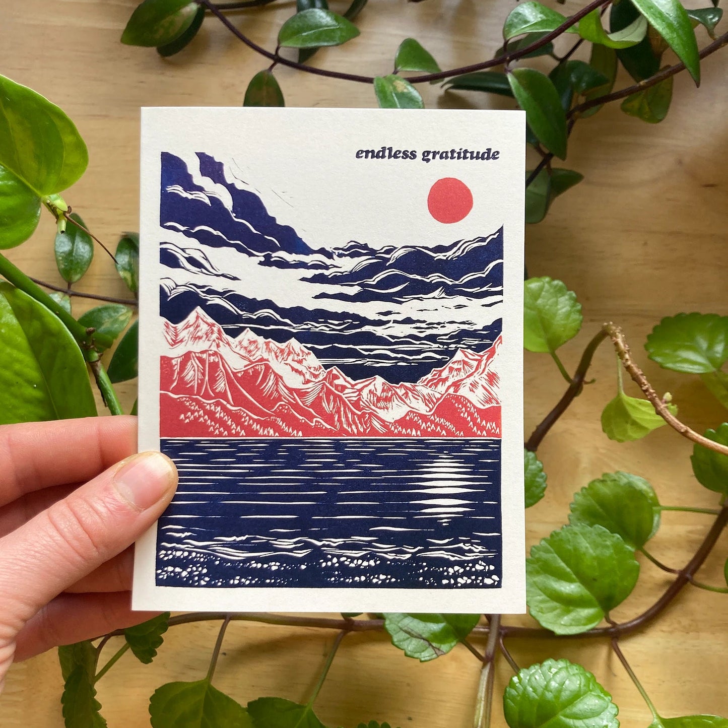 A hand holding a greeting card with a mountain and water artwork, titled 'Endless Gratitude', with a red sun and blue and white mountains, against a wooden background with green leaves.