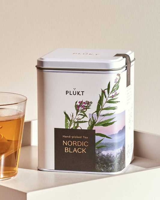 Tea canister labeled 'PLÜkt Nordic Black' with a glass of tea on a light surface.