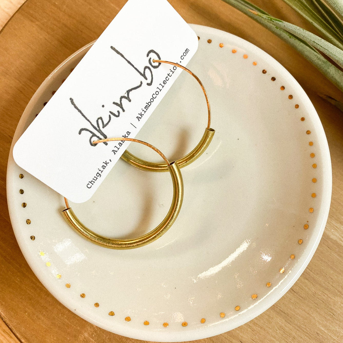 Gold hoop earrings on a white dish with a brand card, placed on a wooden surface with a plant in the background.
