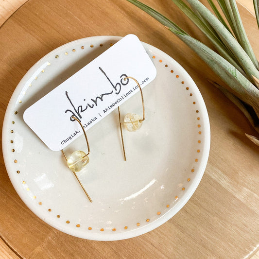 Gold hoop earrings on a white dish with 'akimbo' branding, placed on a wooden surface with a plant.