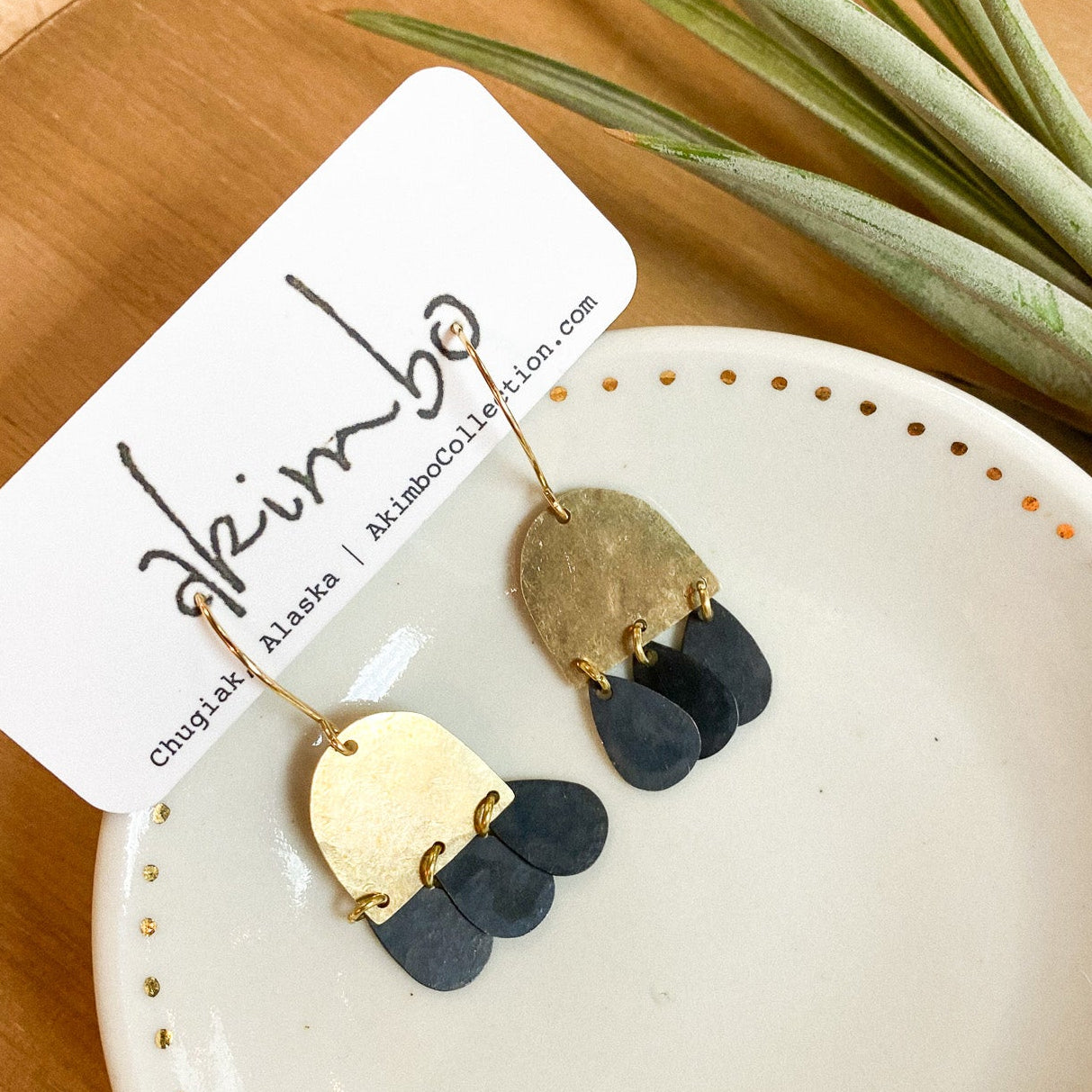 Gold and black earrings on a white dish with a brand tag, placed on a wooden surface with a plant in the background.