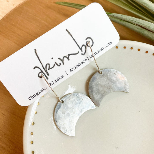 Silver earrings in the shape of a crescent moon on a white dish with a plant in the background