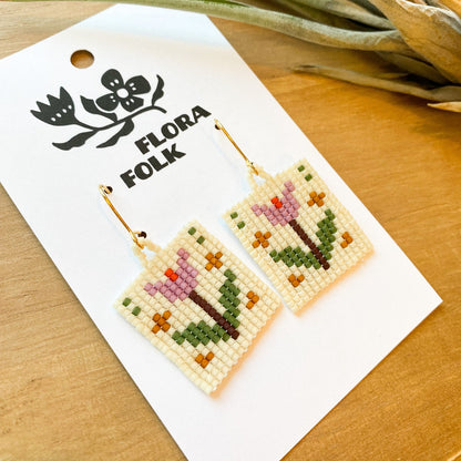Floral beaded earrings on a 'Flora Folk' branded card with a plant in the background.