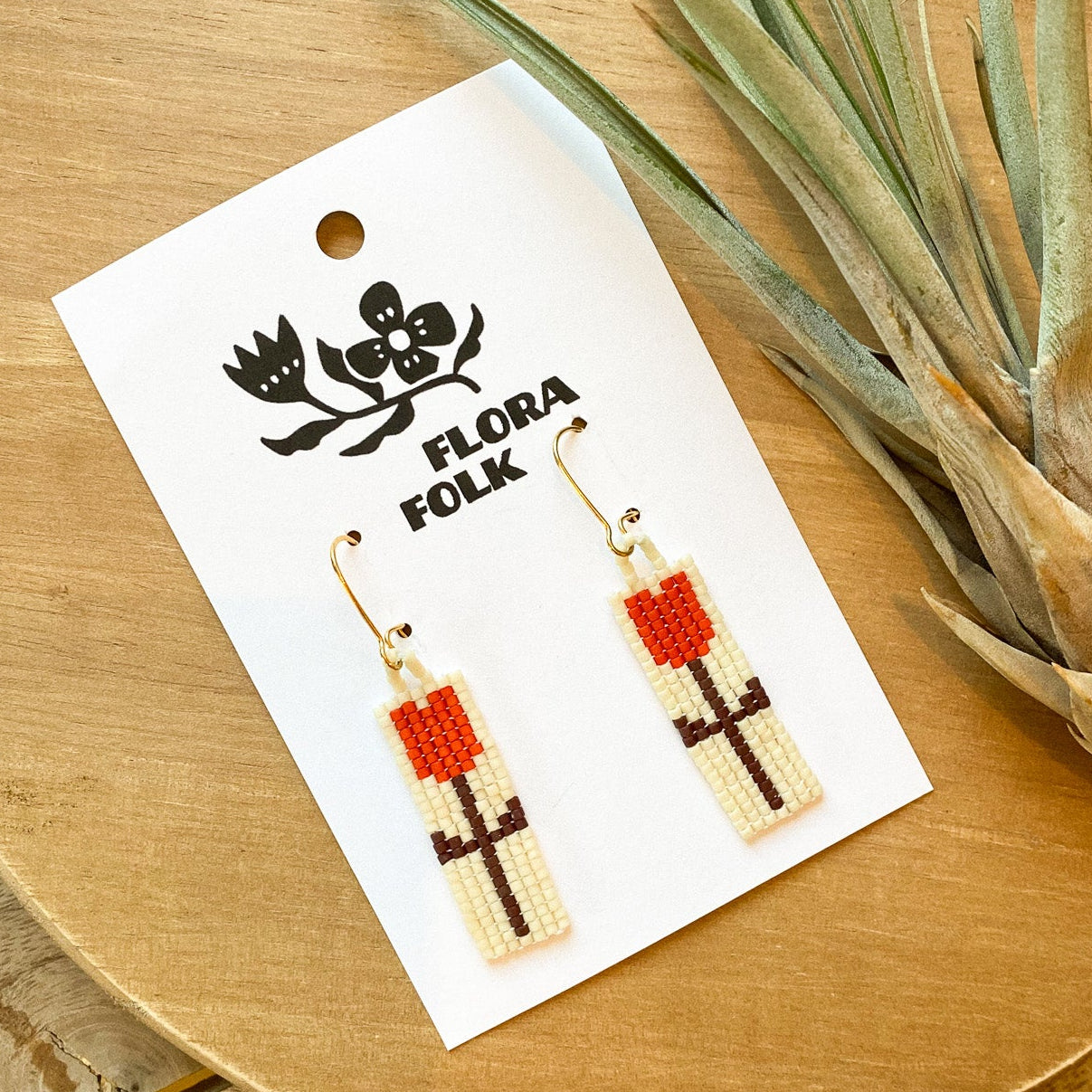 Earrings on a 'Flora Folk' branded card with a plant on a wooden surface