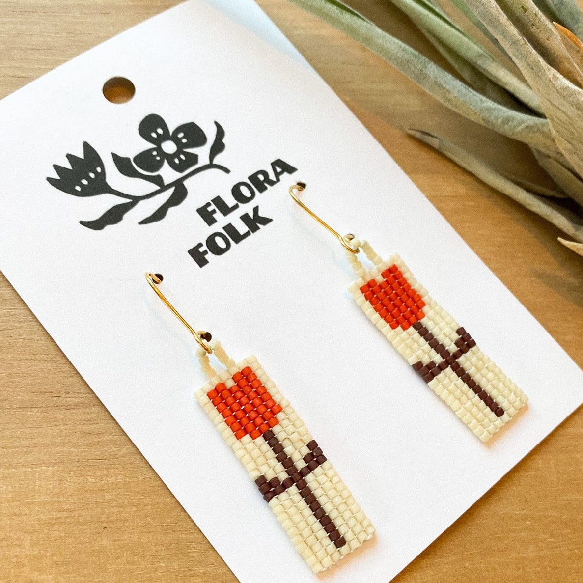 Floral patterned earrings on a 'Flora Folk' branded card with a plant in the background.