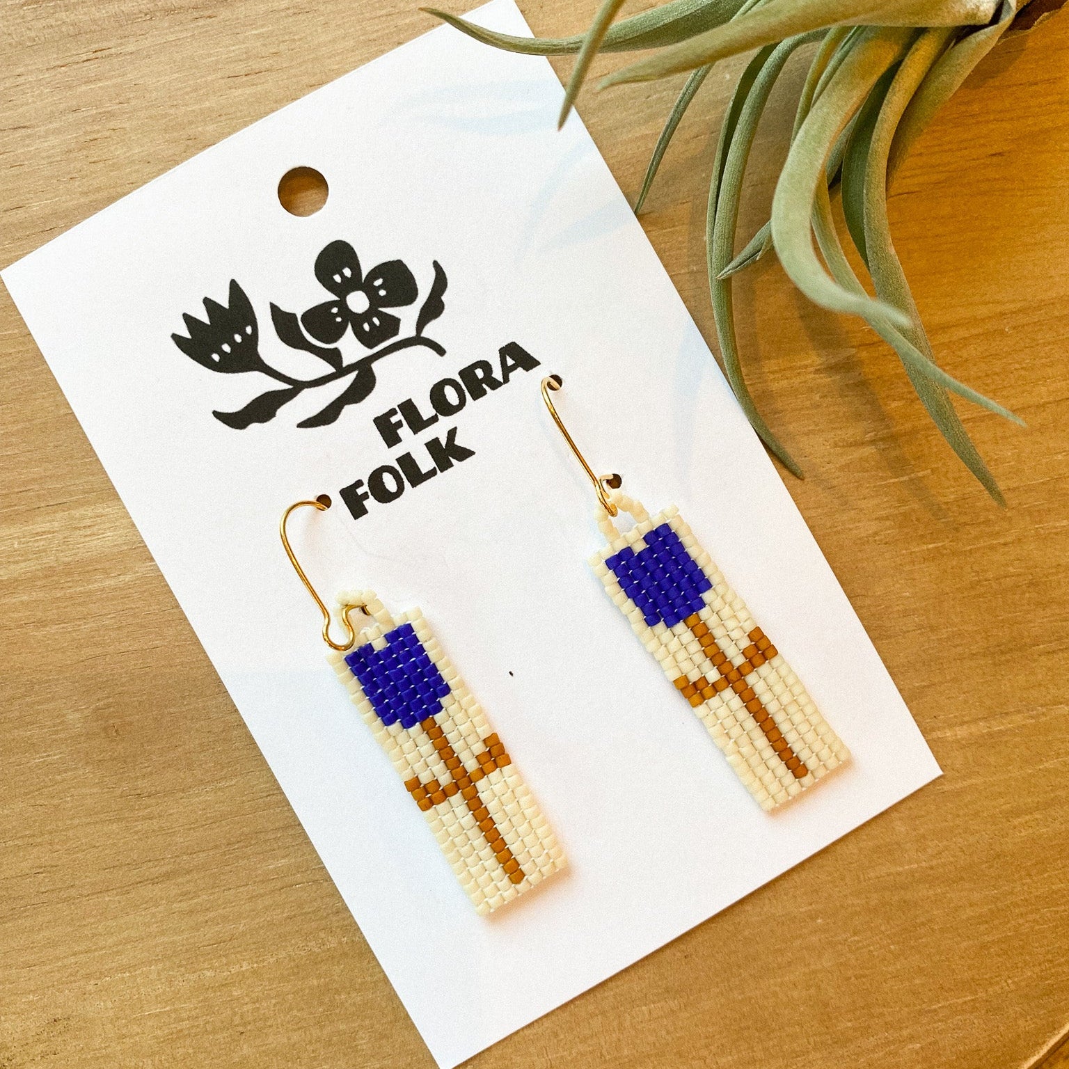 Beaded earrings on a 'Flora Folk' branded card with a plant in the background.