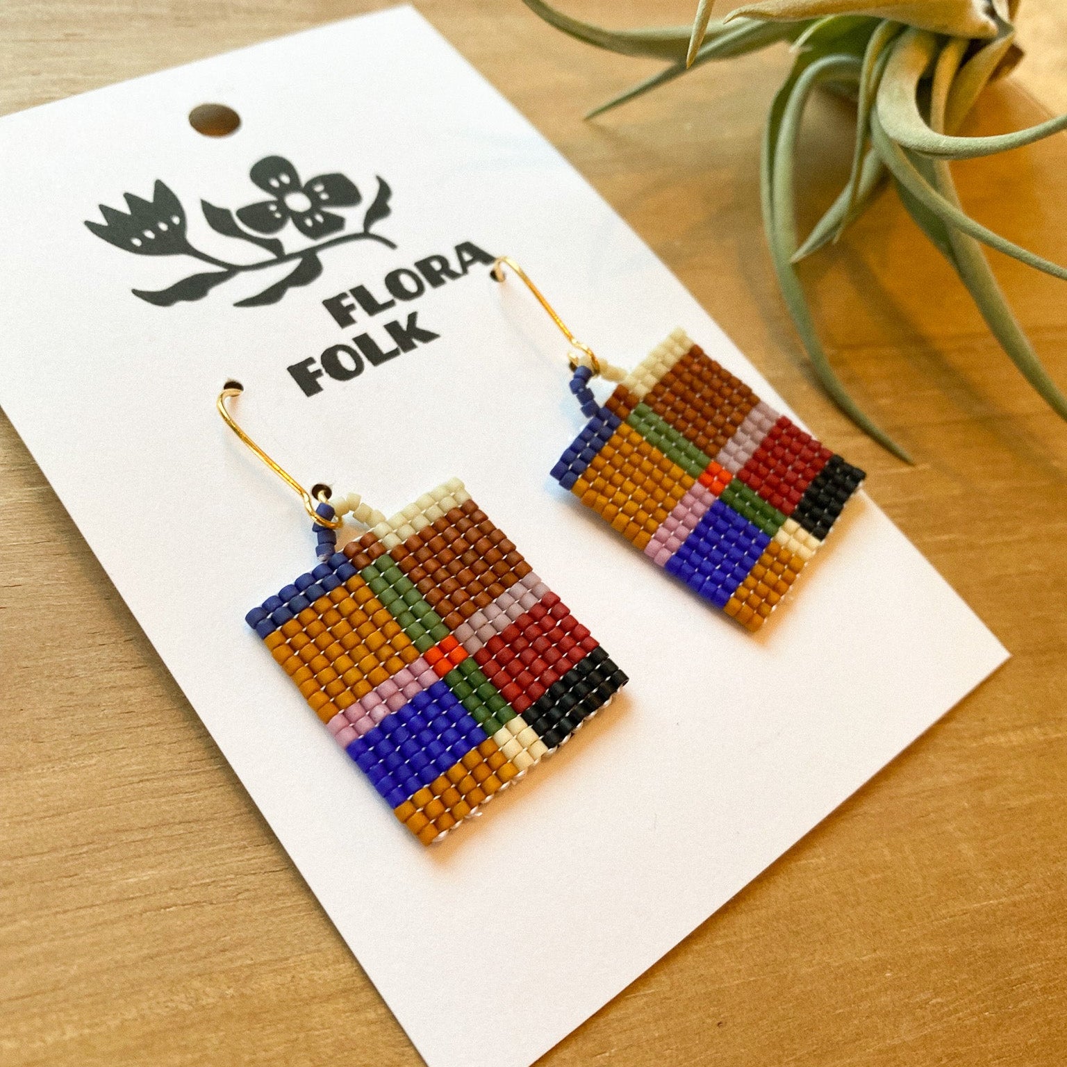 Colorful beaded earrings on a 'Flora Folk' branded card with a plant in the background.