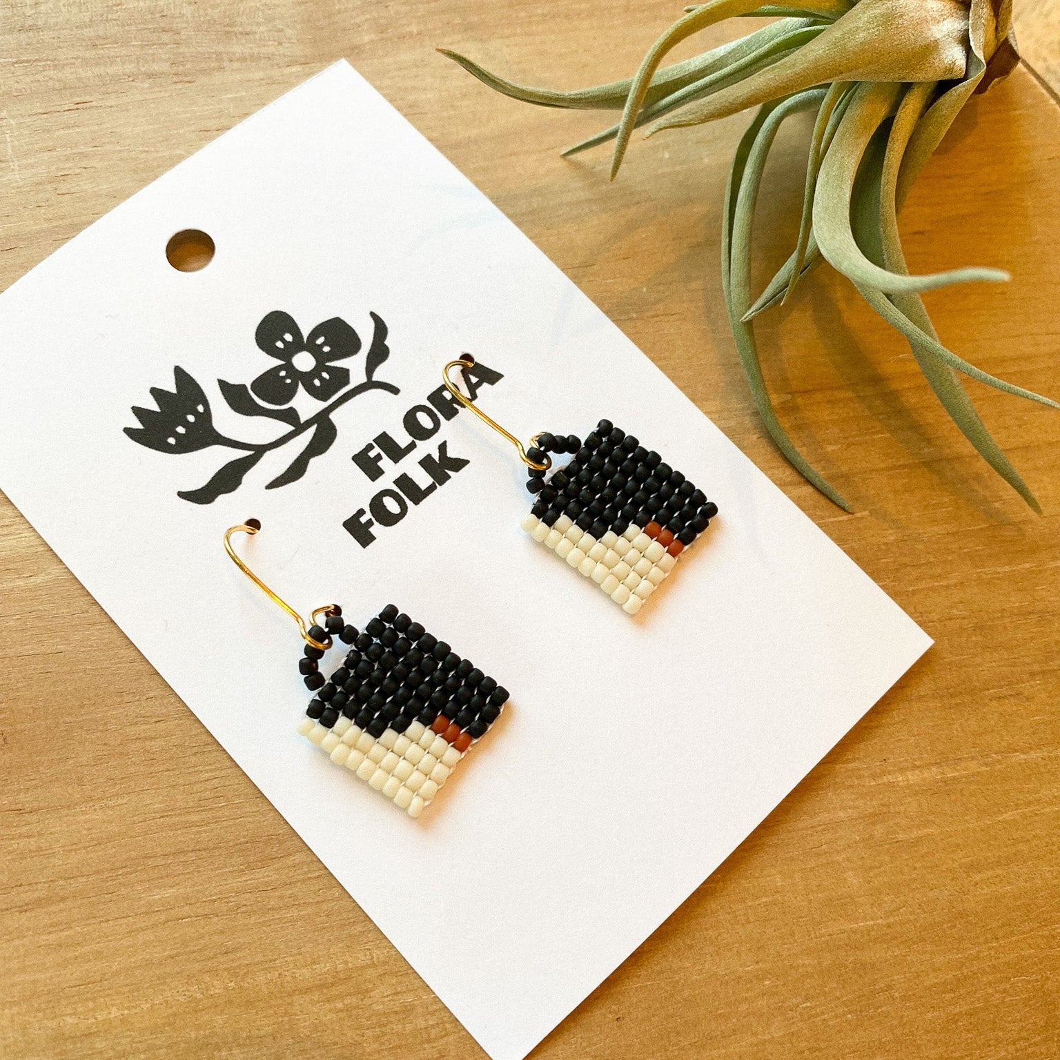 Pair of beaded earrings on a 'Flora Folk' branded card with a plant in the background.