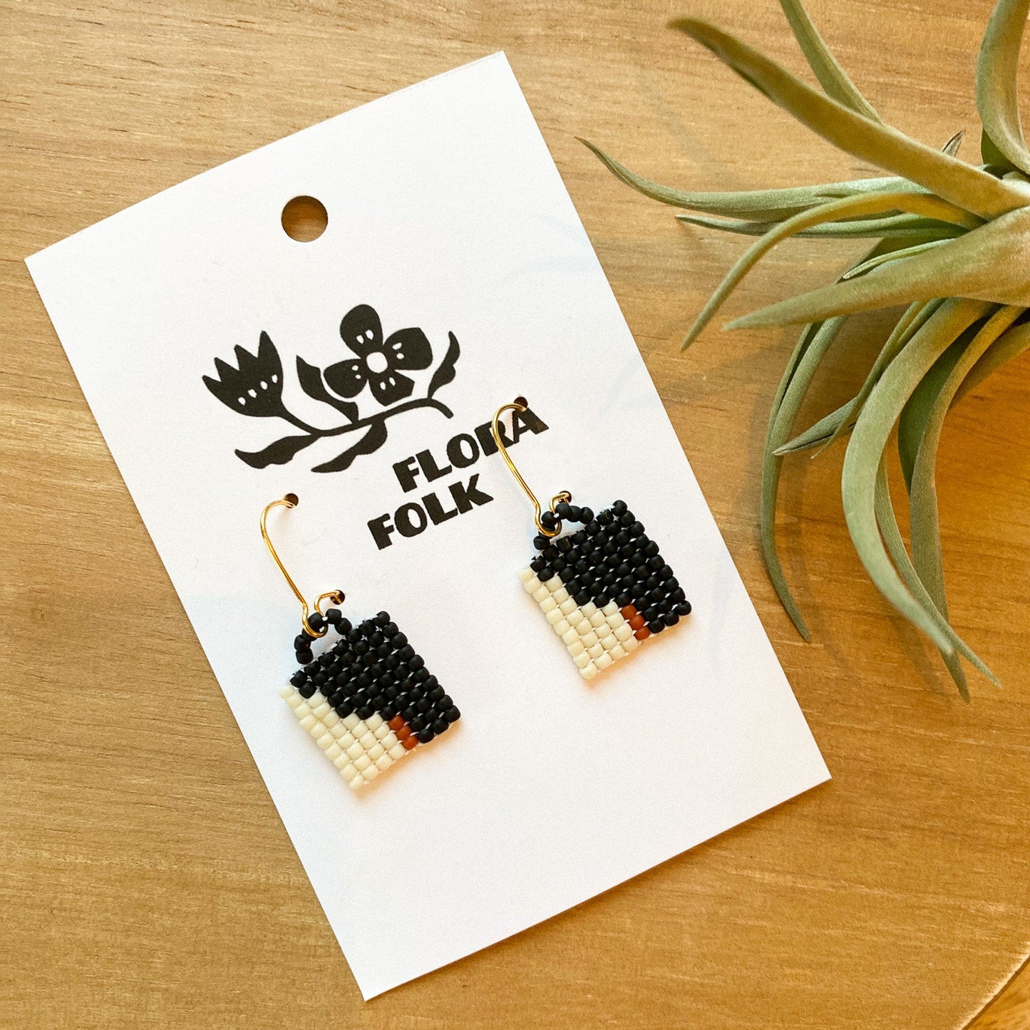 Black and white beaded earrings on a 'Flora Folk' branded card with a plant in the background.