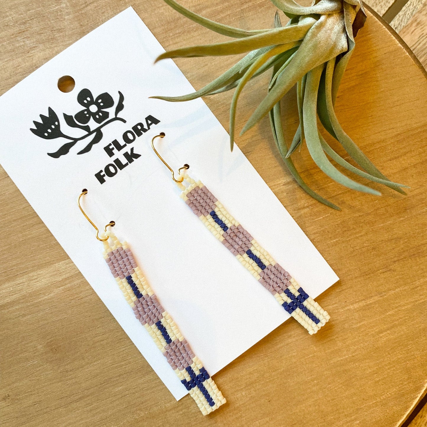 Pair of beaded earrings on a 'Flora Folk' branded card with a plant in the background.