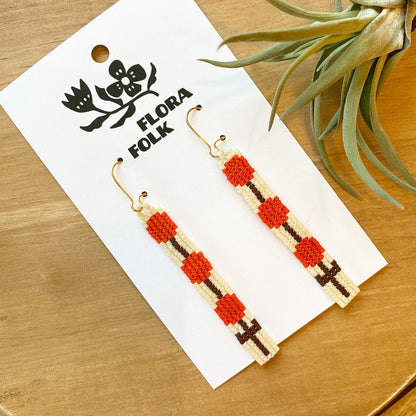 Flora Folk earrings on a wooden surface with a plant in the background