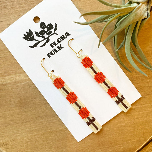 Flora Folk earrings on a wooden surface with a plant in the background