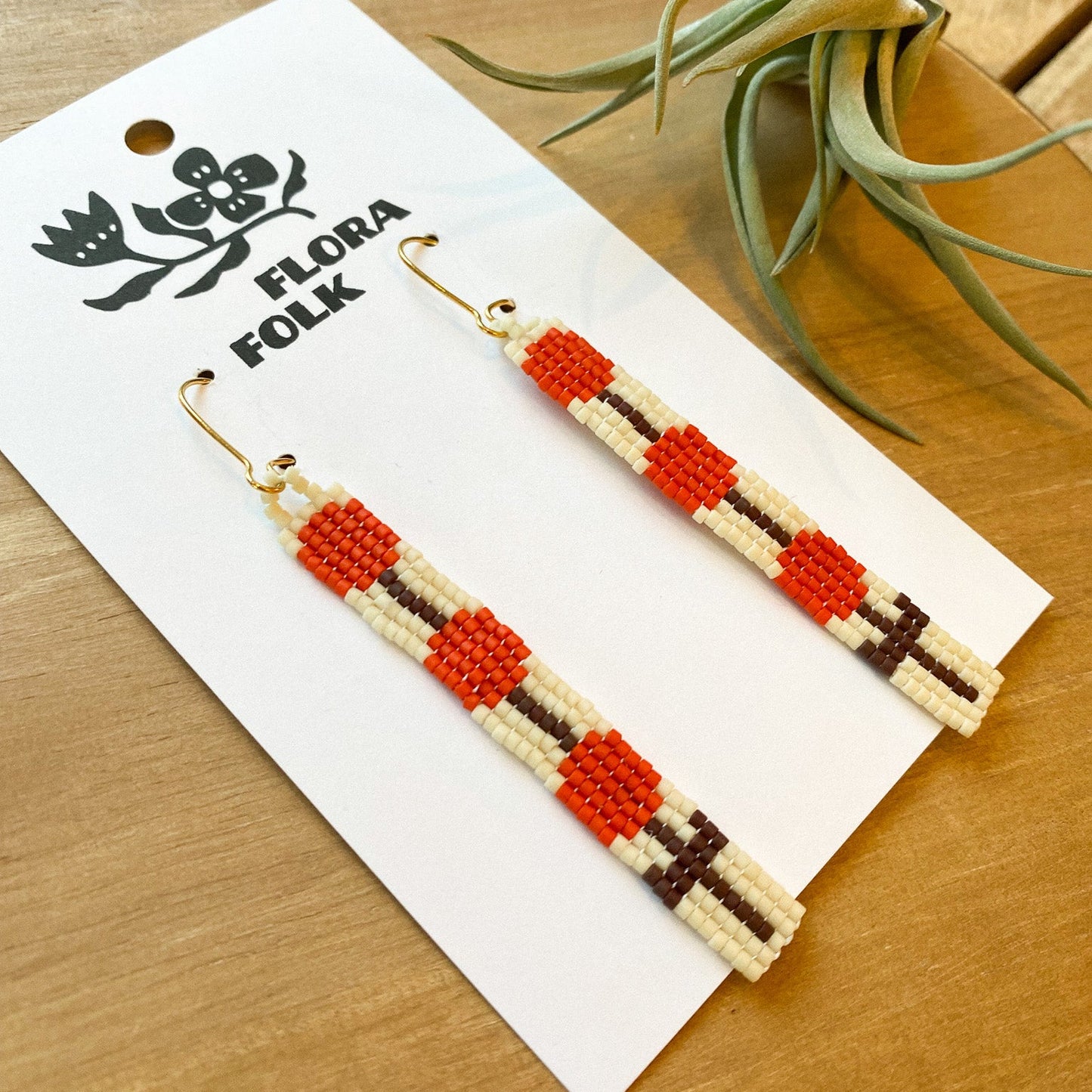 Beaded earrings on a 'Flora Folk' branded card with a plant in the background.