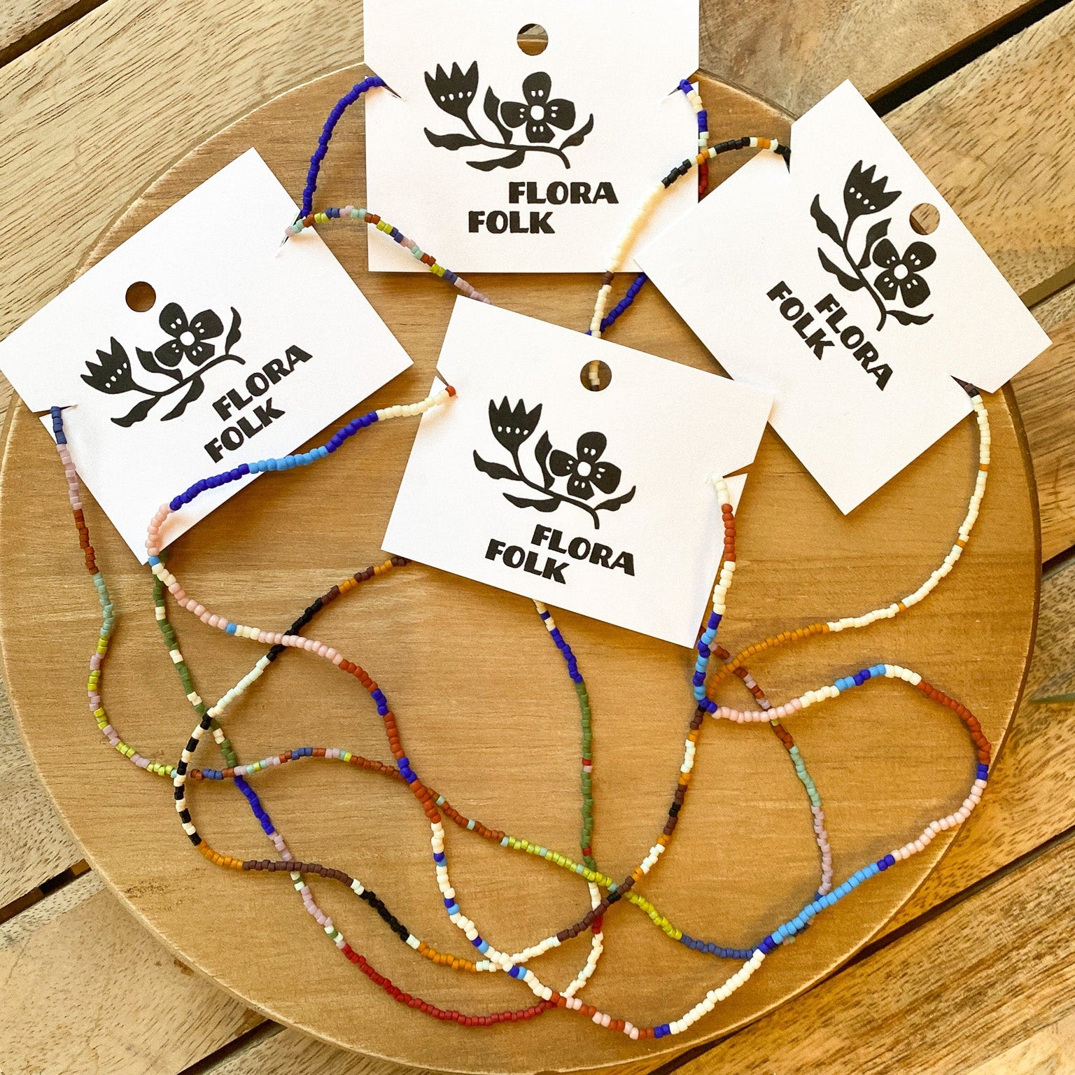 Colorful beaded bracelets on a wooden board with 'Flora Folk' tags.