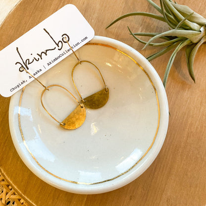 Gold earrings on a white dish with a 'akimbo' tag, placed on a wooden surface with a plant.