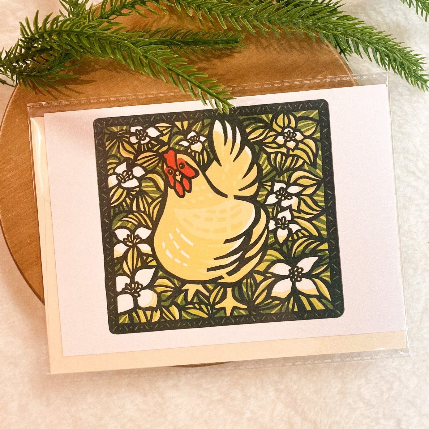 Card with a decorative chicken design on a wooden surface with greenery.