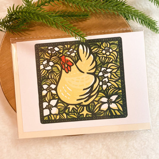 Card with a decorative chicken design on a wooden surface with greenery.
