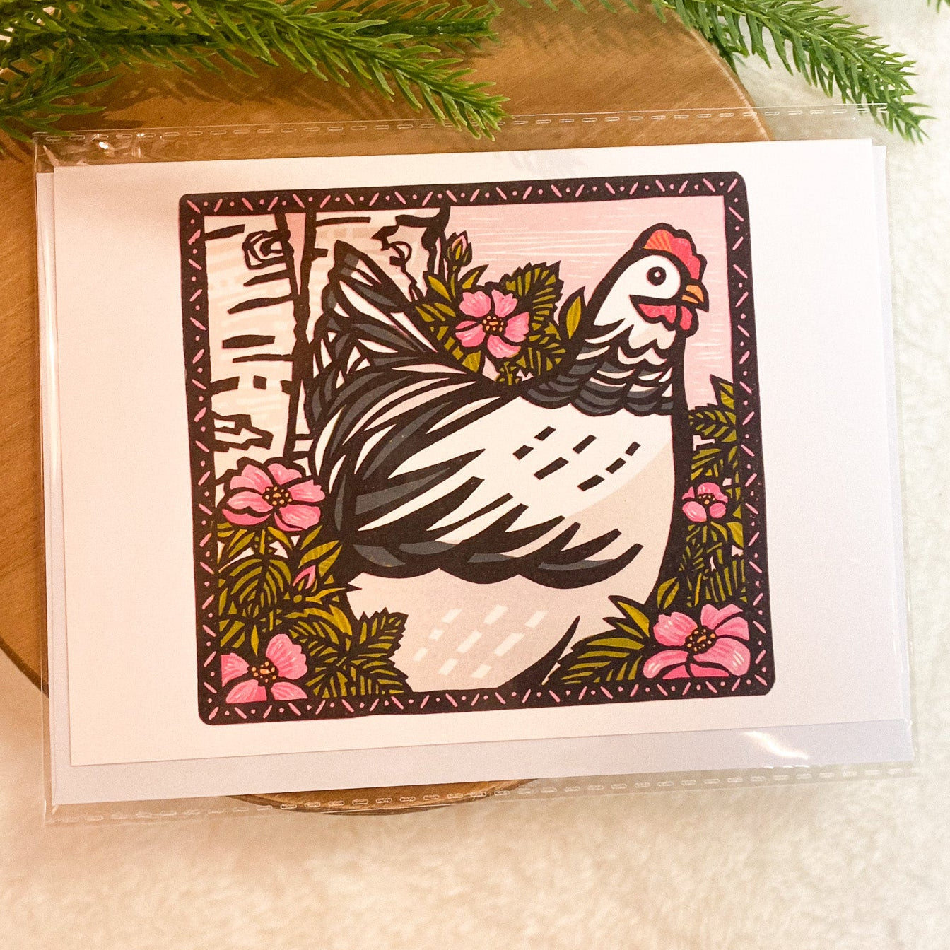 Greeting card with a chicken illustration on a wooden surface with greenery