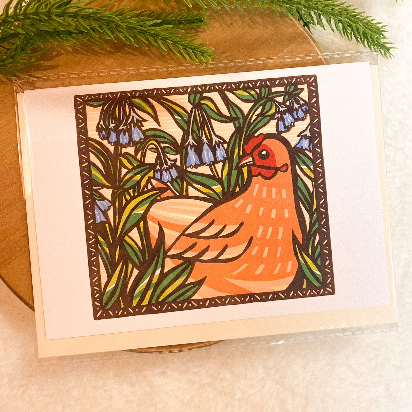 Card with a chicken illustration on a wooden surface with greenery