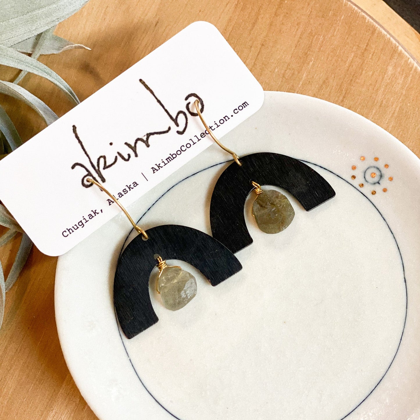 Black and gold earrings on a white dish with 'Akimbo' branding.