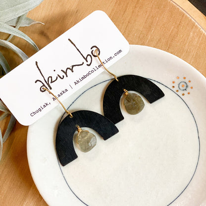Black and gold earrings on a white dish with 'Akimbo' branding.