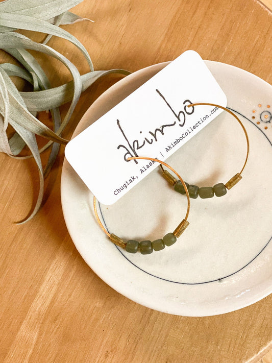 Pair of gold hoop earrings with green beads on a white plate, branded 'Akimbo', on a wooden surface.