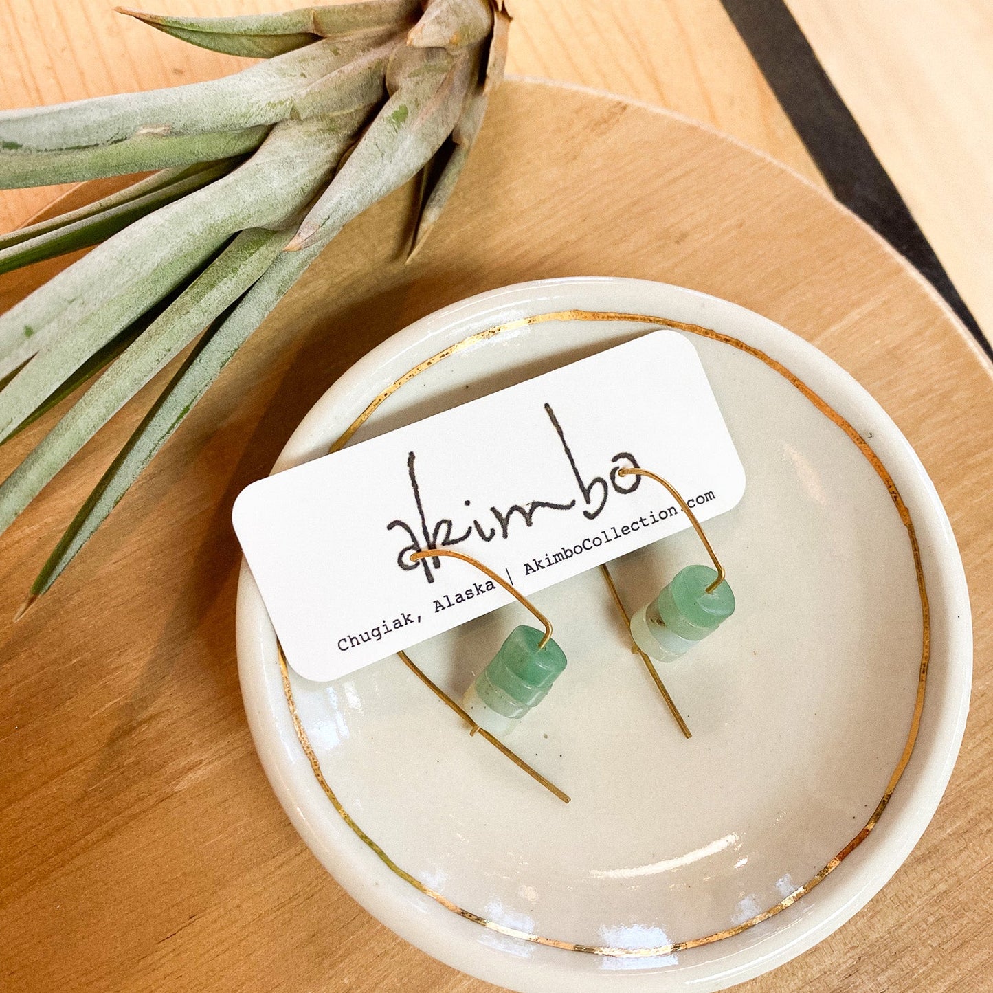 Pair of earrings on a white dish with 'akimbo' label, next to an air plant on a wooden surface.