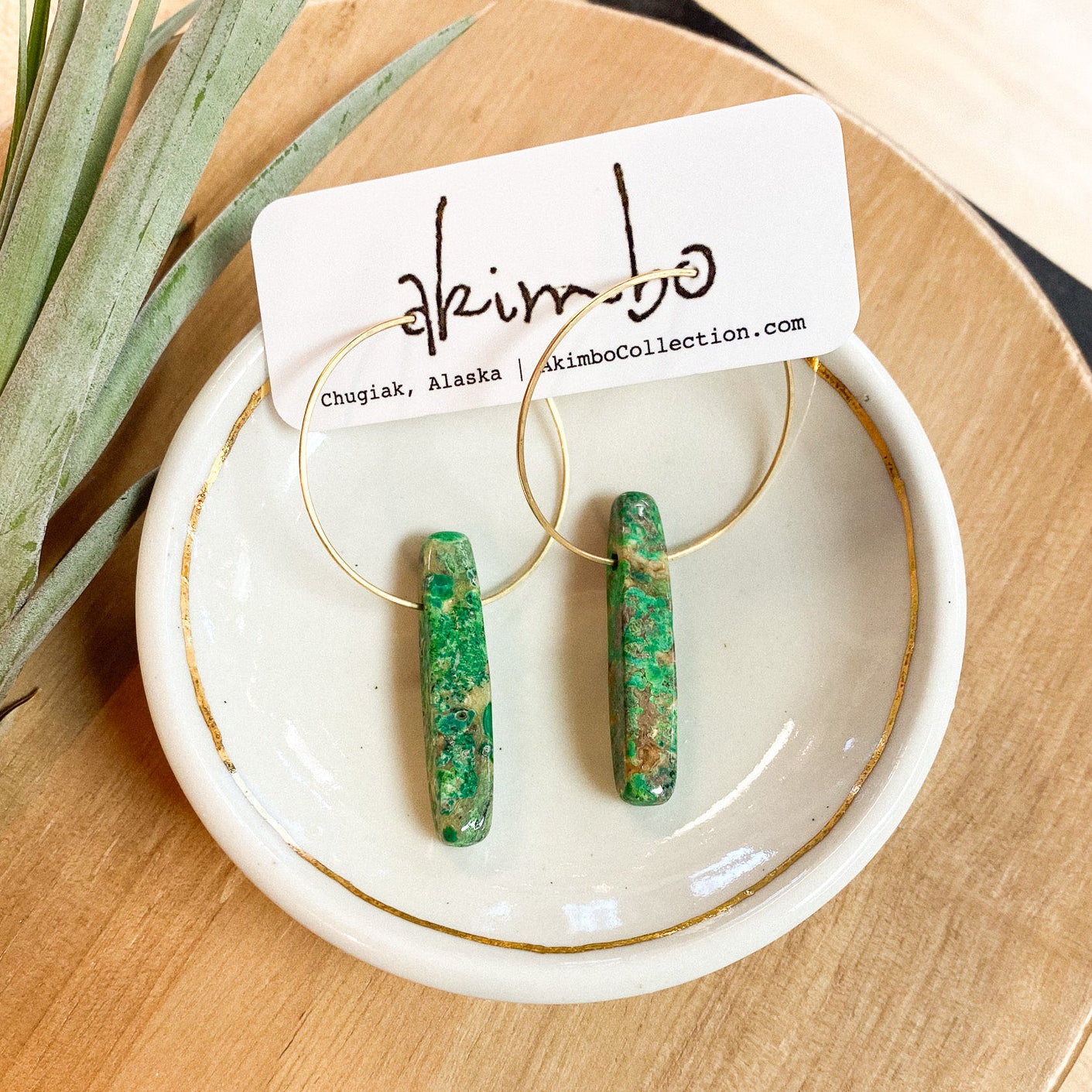 Green earrings on a white dish with 'akimbo' branding on a wooden surface.
