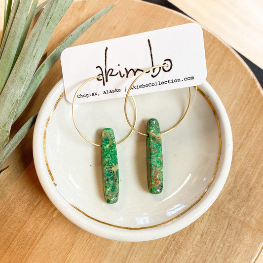 Green earrings on a white dish with 'akimbo' branding on a wooden surface.