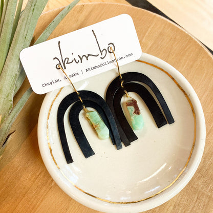 Pair of black and green earrings on a white dish with a 'akimbo' card.