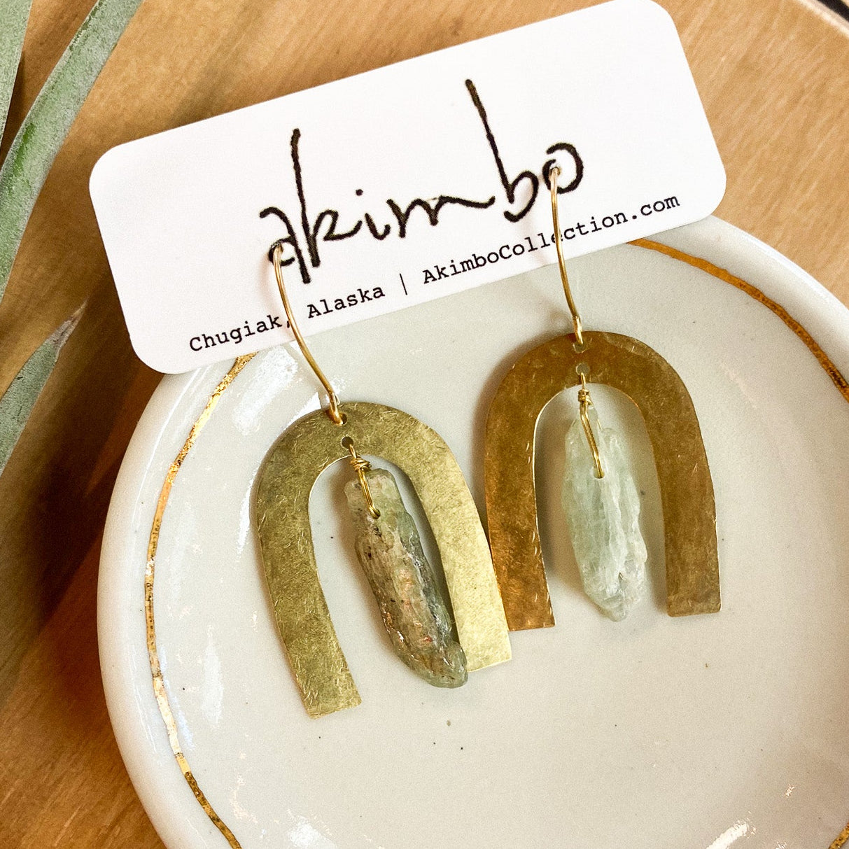 Gold and green arch-shaped earrings on a white dish with 'akimbo' branding.