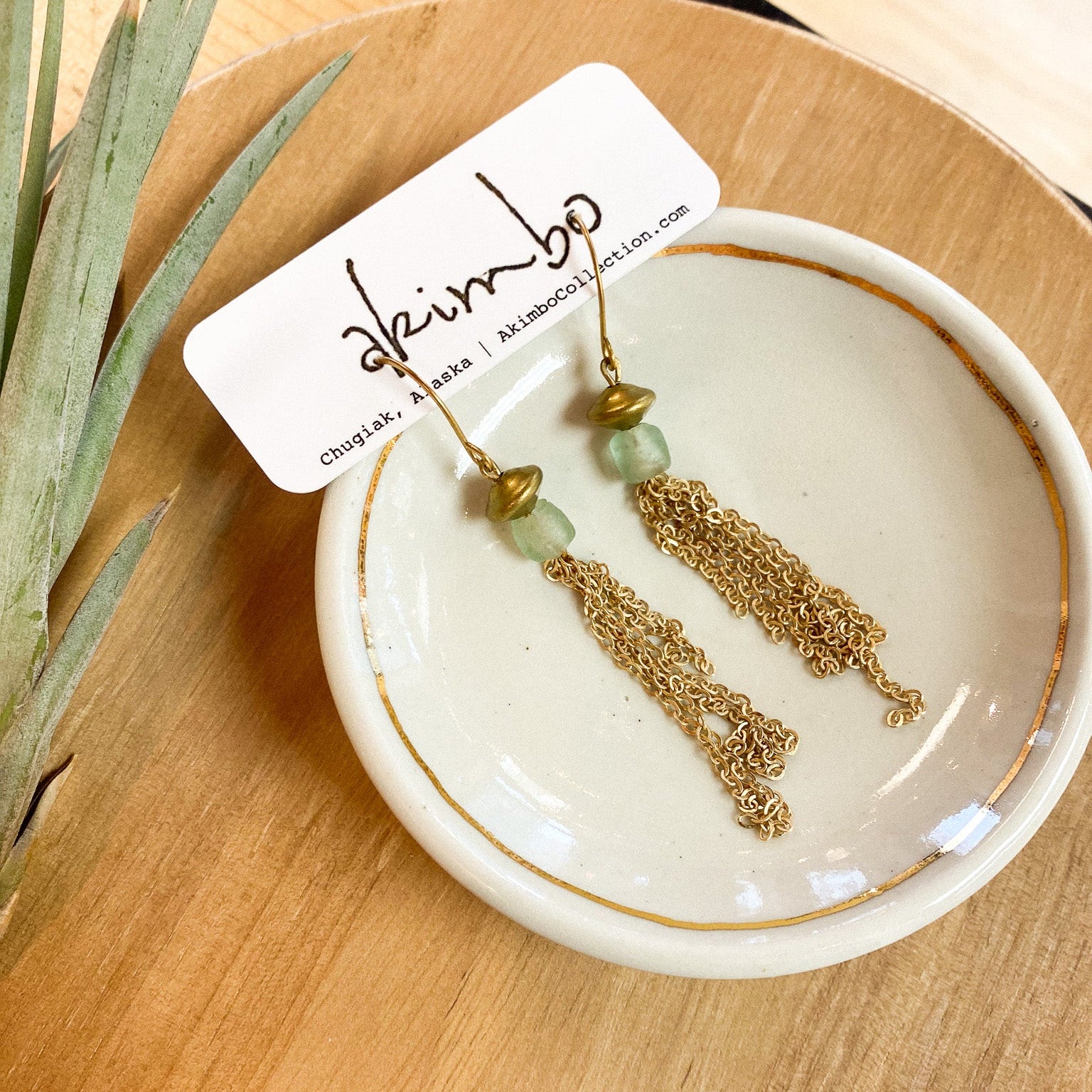 Gold earrings with green beads on a white dish, placed on a wooden surface with a plant in the corner.