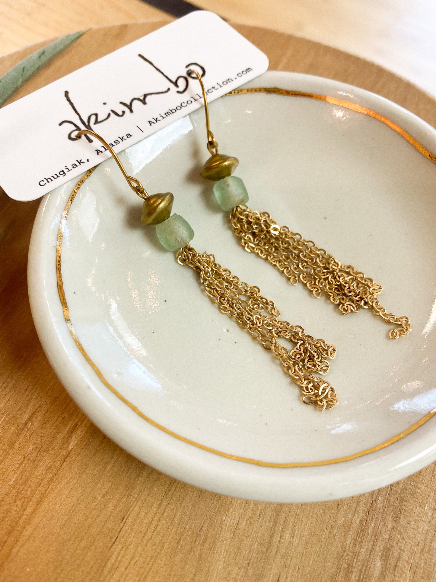 Gold chain earrings with green beads on a white dish, branded 'Akimbo'.