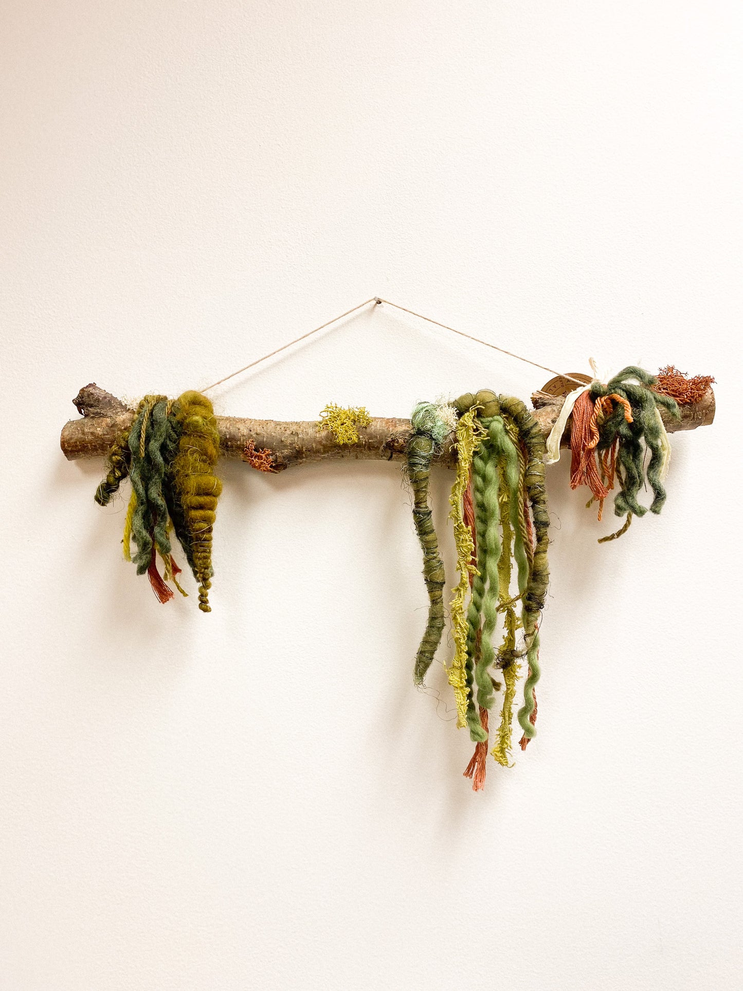 Decorative wall hanging made of a branch with knotted green and brown yarn on a white background